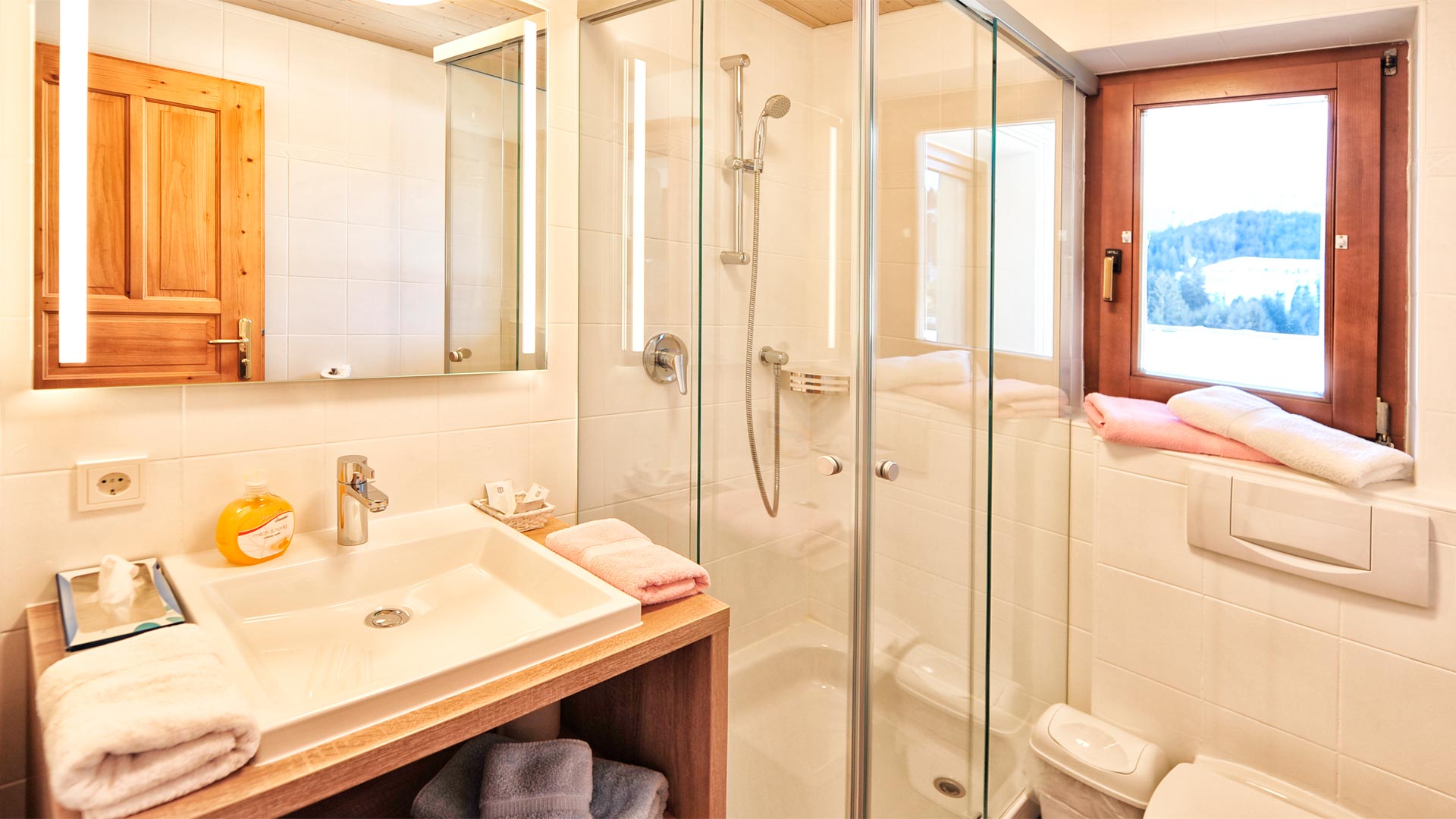 Helles Badezimmer mit Glasdusche, Spiegel, Waschbecken und Fenster mit Blick in verschneite Landschaft, dekoriert mit Handtüchern.
