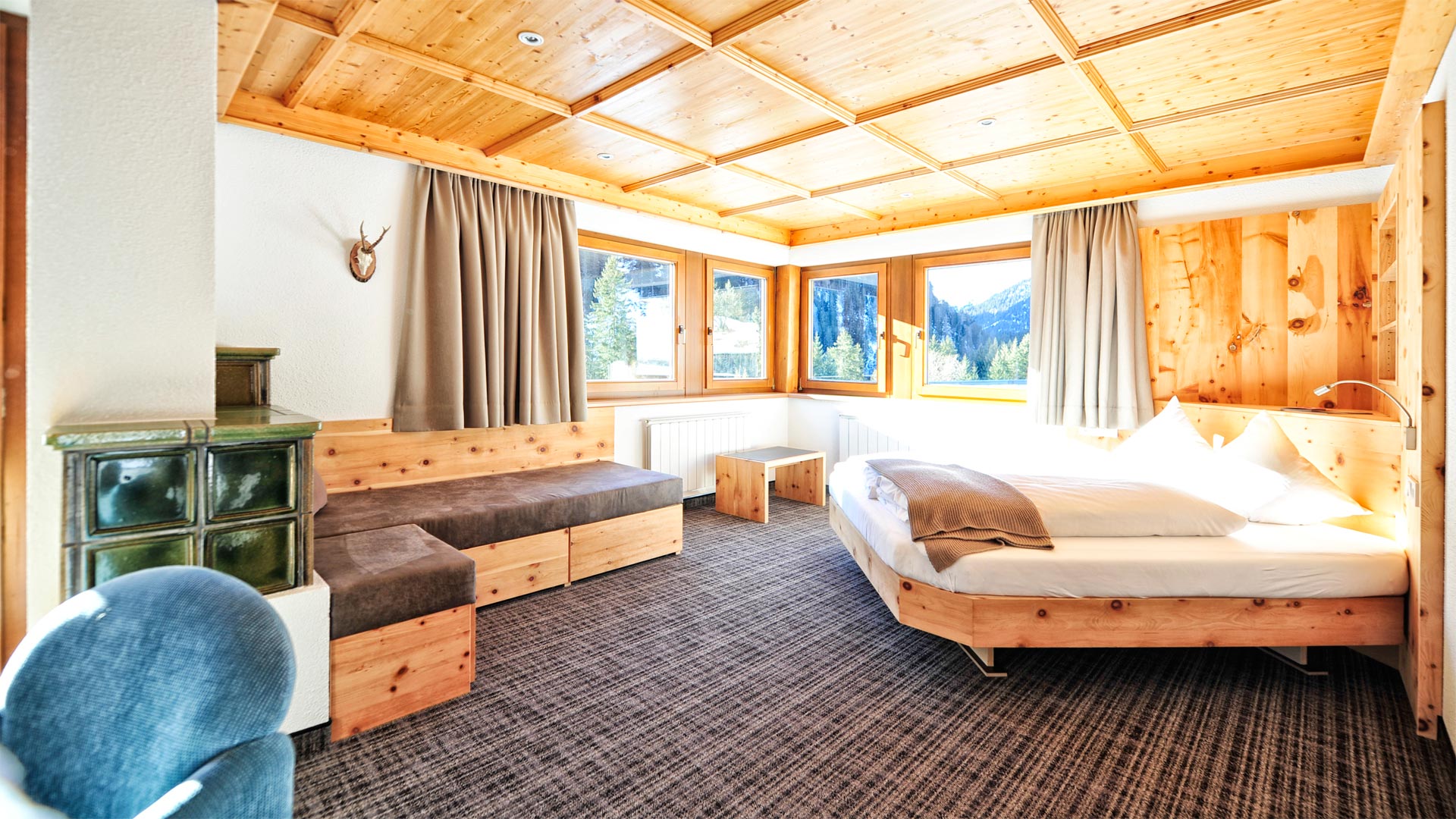 Helles Zirbenholz-Zimmer mit Kachelofen, Bett und Panoramafenstern mit Ausblick auf verschneiten Wald und Berge.