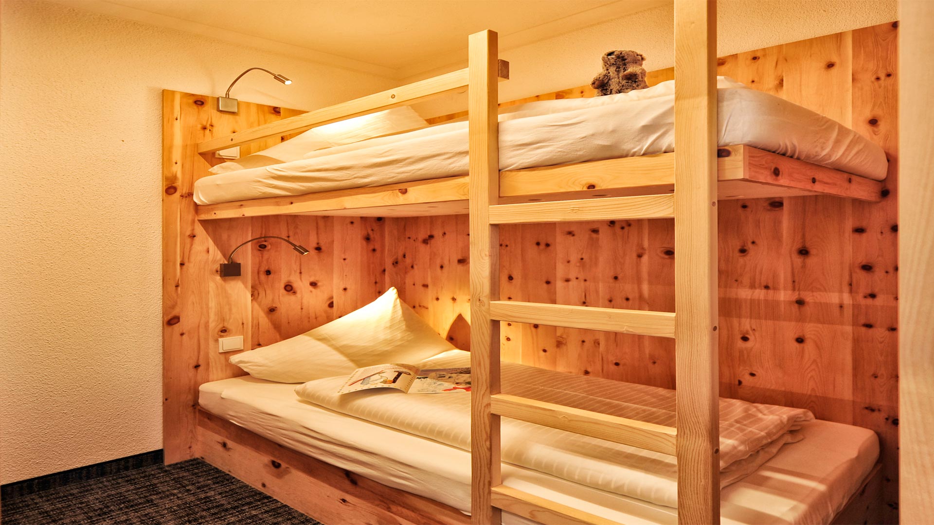 Gemütliches Zimmer mit Etagenbett aus Holz, Leselampen, Kuscheltier und Kinderbuch – ideal für Kinder eingerichtet.