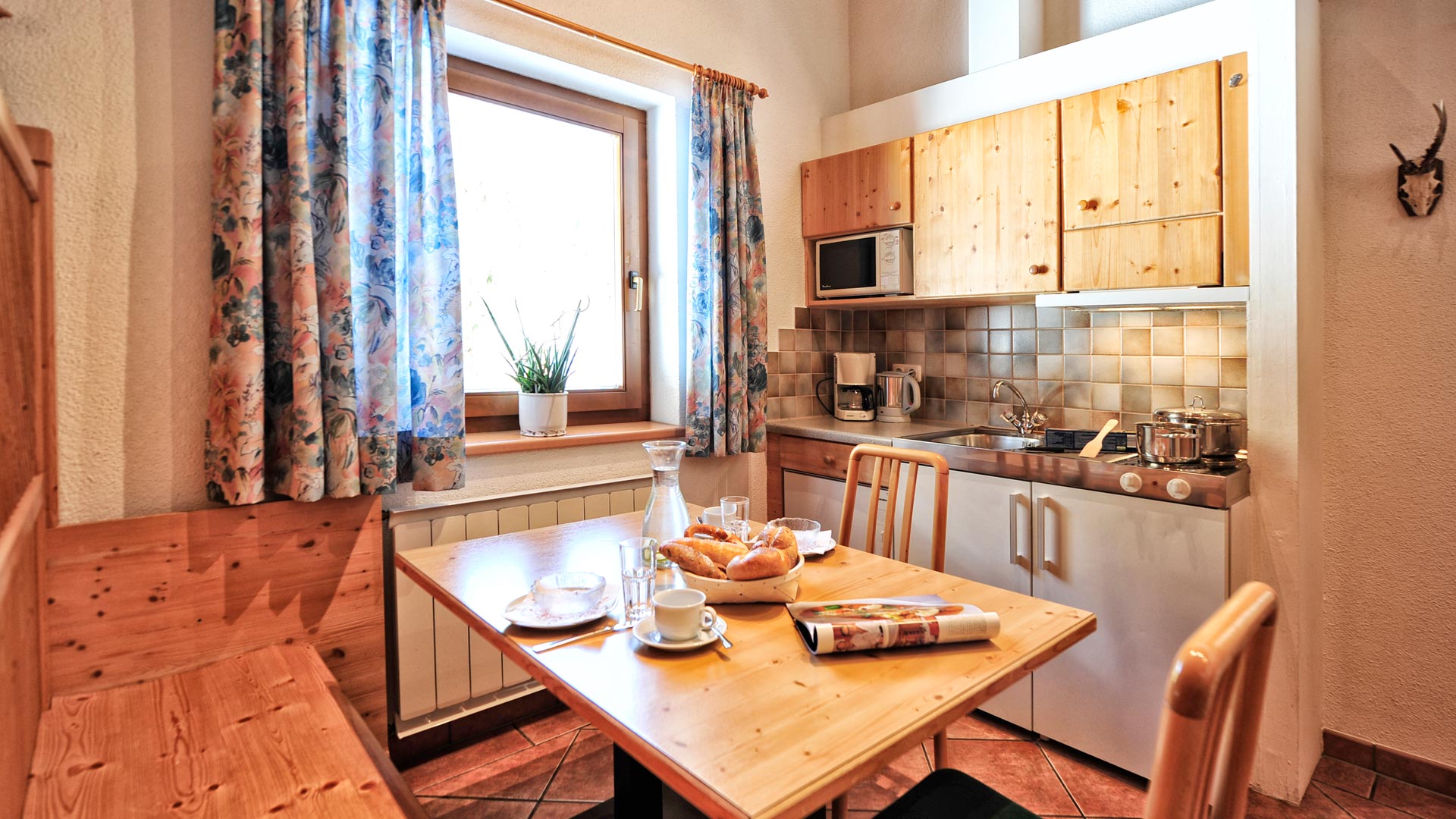 Gemütliche Ferienküche mit Holzmöbeln, Frühstück auf dem Tisch, Kaffeemaschine, Herd und Fenster mit Blumenvorhängen.