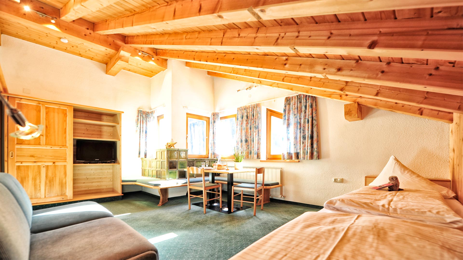 Gemütliches Dachzimmer mit Holzbalkendecke, Einzelbett, Sitzecke, TV, grüner Ofenbank und Fenstern mit Vorhängen.